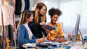 Three people collaborate in a fashion studio, discussing design on a computer. Fabric samples and clothing racks are visible in the background.