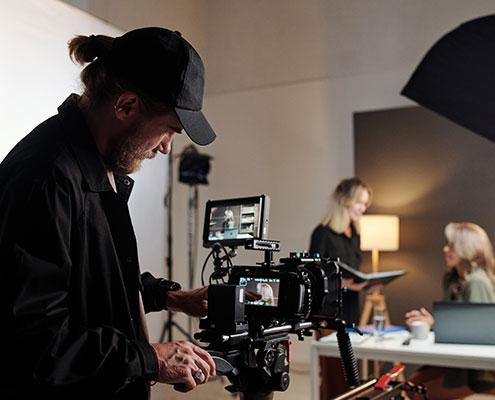 A person operates a camera on a set, focusing on two others discussing at a table, with studio lighting equipment around them.
