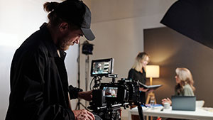A person operates a camera on a film set, capturing two others interacting at a table under studio lighting in a neutral space.