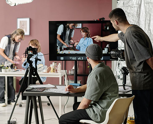A person directs a video production with a crew, cameras, and a monitor displaying the ongoing scene in a studio setting.
