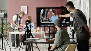 People filming a scene in a studio, with cameras and monitors capturing the action. One person directs while others adjust equipment and prepare.