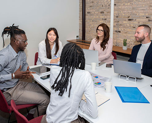 Five people are having a meeting around a table, using laptops and papers. They're in a modern office with a brick wall.