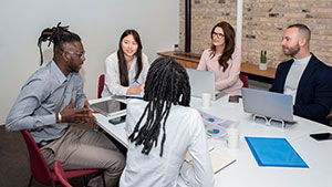 A group of people engaged in a meeting around a table with laptops, papers, and a brick wall background. No landmarks visible.