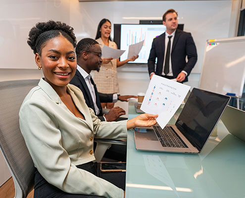 A group of professionals in a meeting room discussing charts and graphs, with one person smiling while holding a paper near a laptop.
