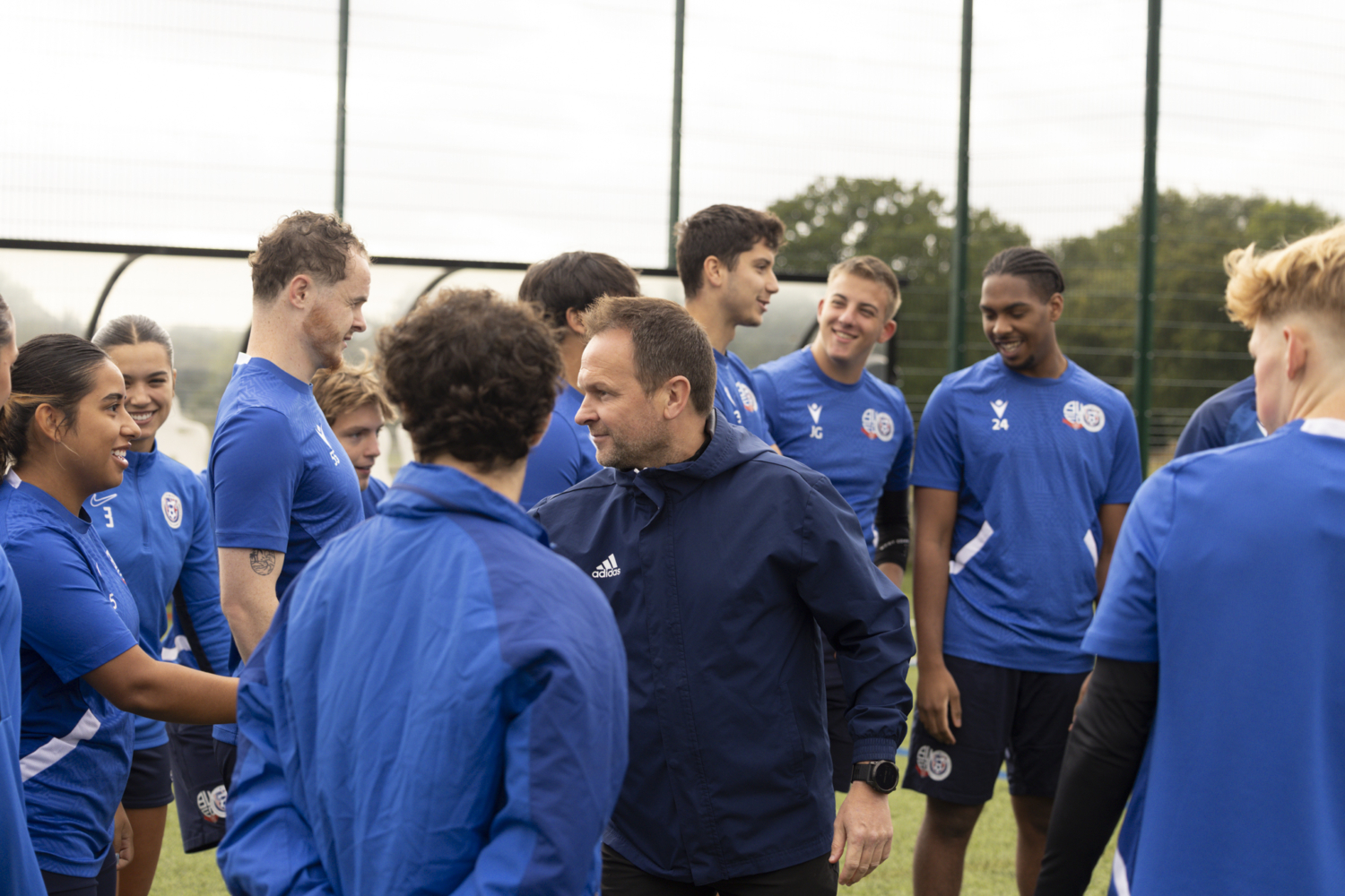 A group of people wearing blue sports attire gather on a field, engaging in friendly conversation, with trees and a fence in the background.