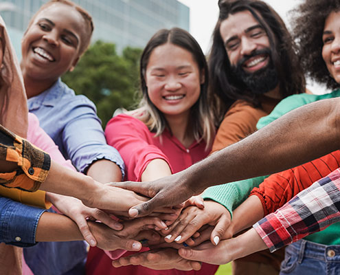 A diverse group of people stand outdoors with hands joined in the center, smiling and radiating unity, against a background of trees and buildings.