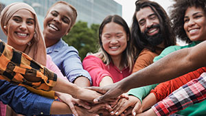 A group of diverse people smiling, standing in a circle with hands joined in the center against an urban backdrop.