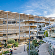 Universite-de-Montpellier,-France A modern, multi-story building with steel and glass facade, surrounded by pathways and greenery. People walk nearby under a clear blue sky.