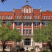University-of-the-Incarnate-Word A historic red-brick building with ornate architectural details, bordered by trees. The foreground features a small statue and a clear blue sky.