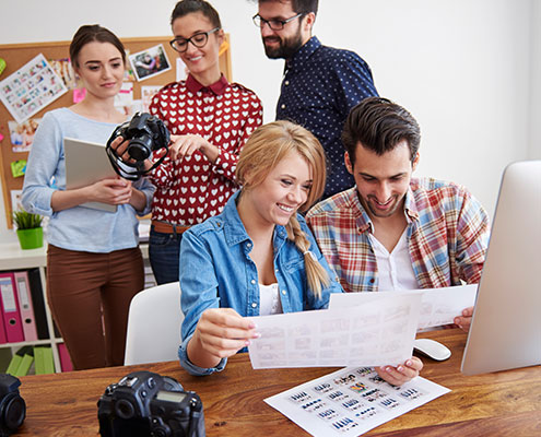 A group of five people in a creative office setting, reviewing photographs and camera equipment, collaborating around a wooden table with enthusiasm.