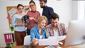 Five people in a casual office environment collaborate on a project, reviewing documents and using a computer, with a bulletin board in the background.