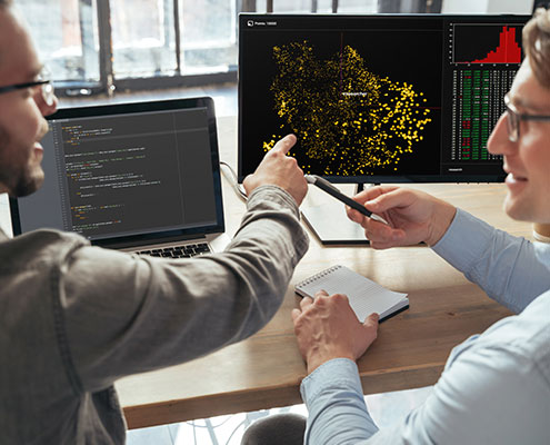 Two people discuss data visualizations on computer screens, pointing at graphs. A notebook and pen lie on the desk in a modern office setting.