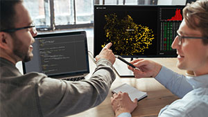 Two people discuss data on computer screens with code and analytics. One points at the screen, while the other holds a pen.
