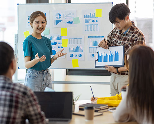 A group of people in a meeting room discuss charts and graphs on a whiteboard and tablet during a presentation.