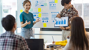A group of people engage in a presentation with charts and graphs on a whiteboard in an office setting, discussing data analysis.