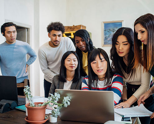 A group of seven people gathered around a laptop in an office setting, engaging in a collaborative discussion with various documents and plants around.