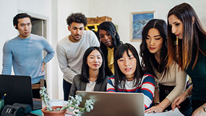 A group of seven people gathers around a laptop in a modern office setting, engaging in a collaborative discussion.