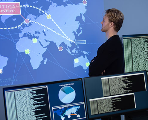A person observes a digital map on a screen, with multiple computer monitors displaying data and charts, indicating cybersecurity or global network analysis.