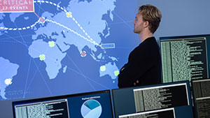 A person stands in front of multiple monitors showing data and a world map, likely analyzing global information or cybersecurity events.