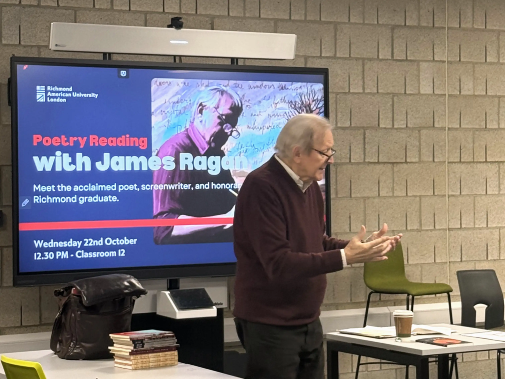James Ragan stands near a digital screen displaying a poetry reading event at Richmond American University London, with books and coffee on a table.