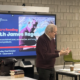 James Ragan stands near a digital screen displaying a poetry reading event at Richmond American University London, with books and coffee on a table.