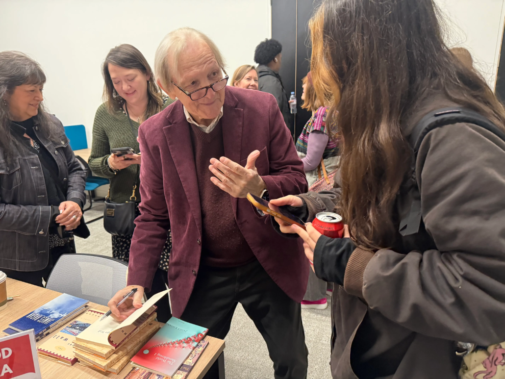 A person in a maroon jacket signs a book for another person, surrounded by others in a casual indoor setting.