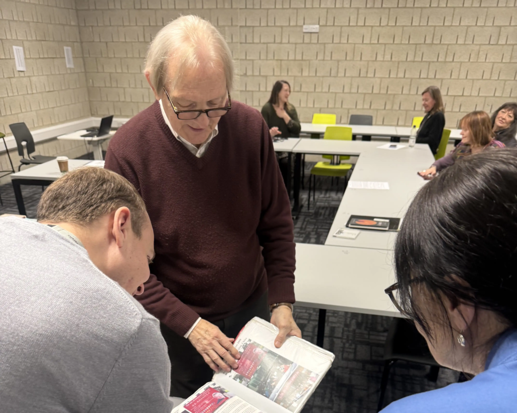 A group of people in a classroom setting, one person showing a page from a book to others seated around tables.