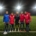 5 people standing on a green football pitch wearing football kits with stadium lights in the background