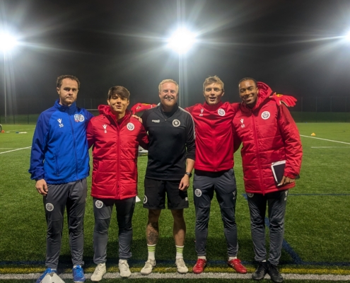 5 people standing on a green football pitch wearing football kits with stadium lights in the background