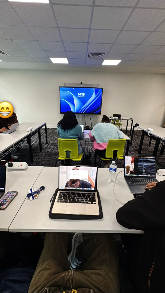 People seated in a classroom, working on laptops. A large screen displays "14:19" at the front. Various devices are on desks.