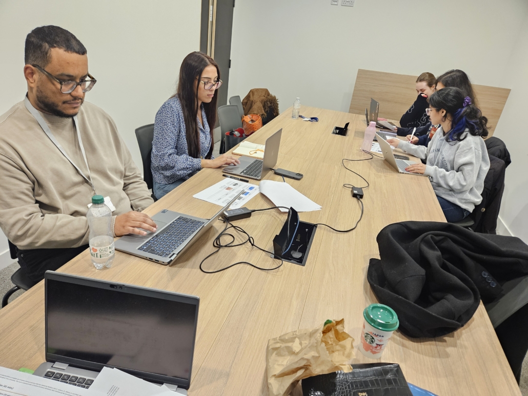 Five people working on laptops at a conference table, engaged in collaboration. Documents, water bottles, and coffee cups are on the table.