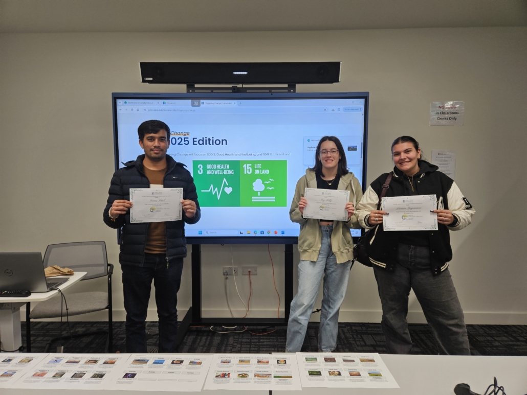 Three people proudly hold certificates in front of a presentation screen displaying "2025 Edition." Classroom setting with chairs, table, and laptop visible.