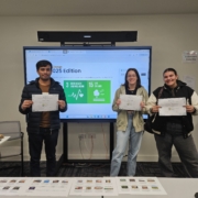 Three people proudly hold certificates in front of a presentation screen displaying "2025 Edition." Classroom setting with chairs, table, and laptop visible.