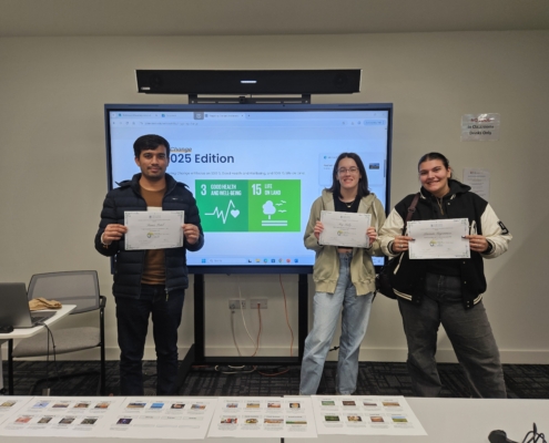 Three people proudly hold certificates in front of a presentation screen displaying "2025 Edition." Classroom setting with chairs, table, and laptop visible.