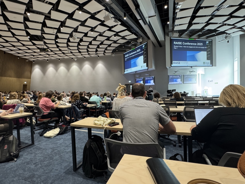 A large conference room filled with numerous people at the "RAISE Conference 2025," featuring a modern design and multiple presentation screens.