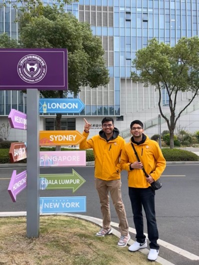 Two people in yellow jackets stand by a colorful directional sign with city names. A modern glass building is in the background.
