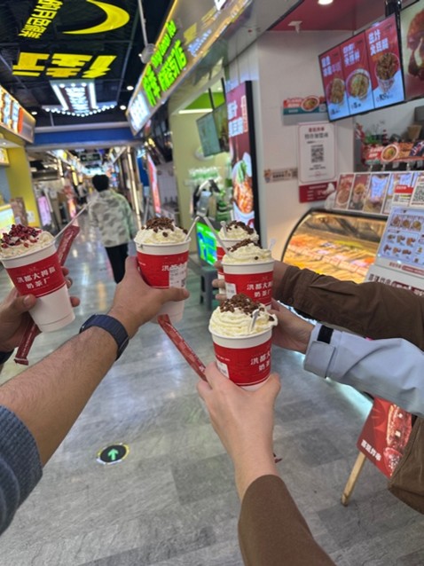 Five people holding up red cups with whipped cream in a busy indoor food market, surrounded by bright food signage and displays.
