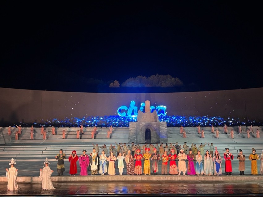 A nighttime stage performance featuring many individuals in traditional attire, with "China" illuminated in the background and a stone structure present.