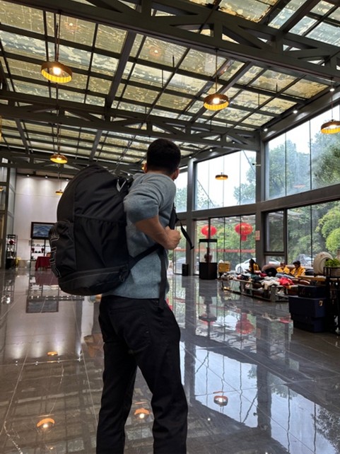 A person with a large backpack stands in a bright, modern hallway with high ceilings, looking toward large windows with outdoor views.