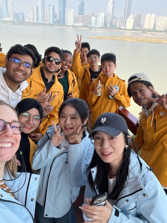 A group of people in yellow and blue jackets posing happily on a boat, with a city skyline visible in the background.