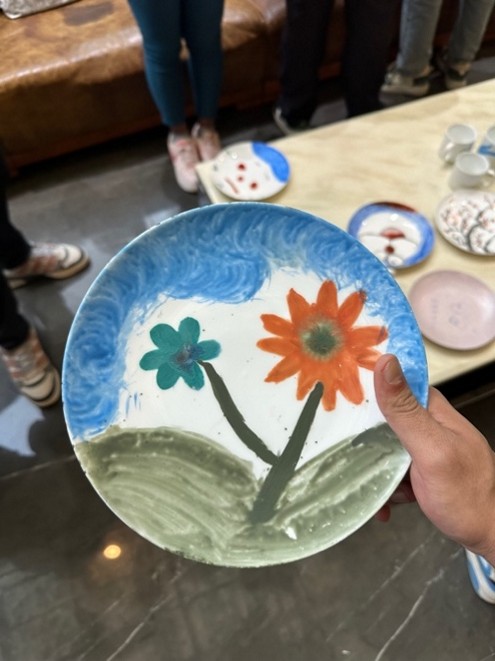 A person holds a hand-painted plate depicting colorful flowers and sky. Other patterned plates and people are visible in the background.