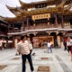 A person stands before the ornate Yu Garden shopping area in Shanghai, with traditional architecture and other people strolling nearby.