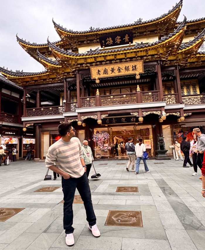 A person stands before the ornate Yu Garden shopping area in Shanghai, with traditional architecture and other people strolling nearby.