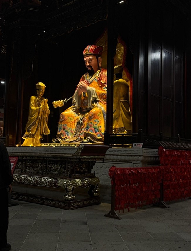 A large, colorful statue in traditional attire sits in a dimly lit temple room, surrounded by a person and decorative elements.