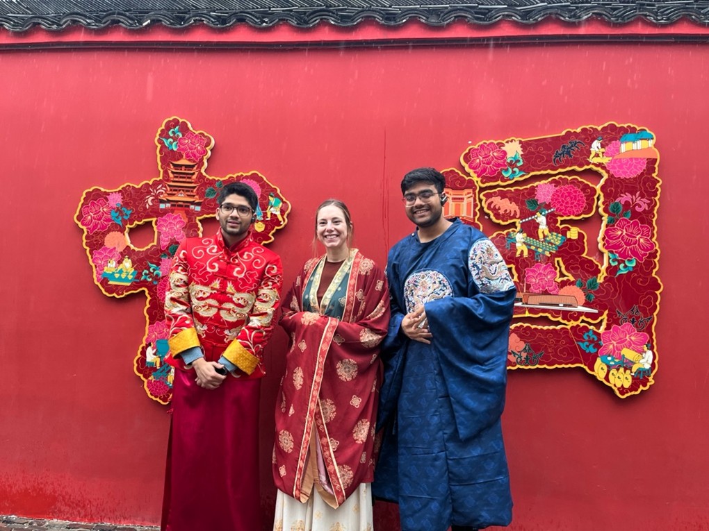 Three people in traditional colorful garments stand against a red wall with decorative Chinese symbols, smiling for a photo.