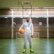 A person stands in a sports hall holding a basketball, wearing a sash. Indoor green court and goals are visible in the background.