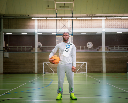 A person stands in a sports hall holding a basketball, wearing a sash. Indoor green court and goals are visible in the background.