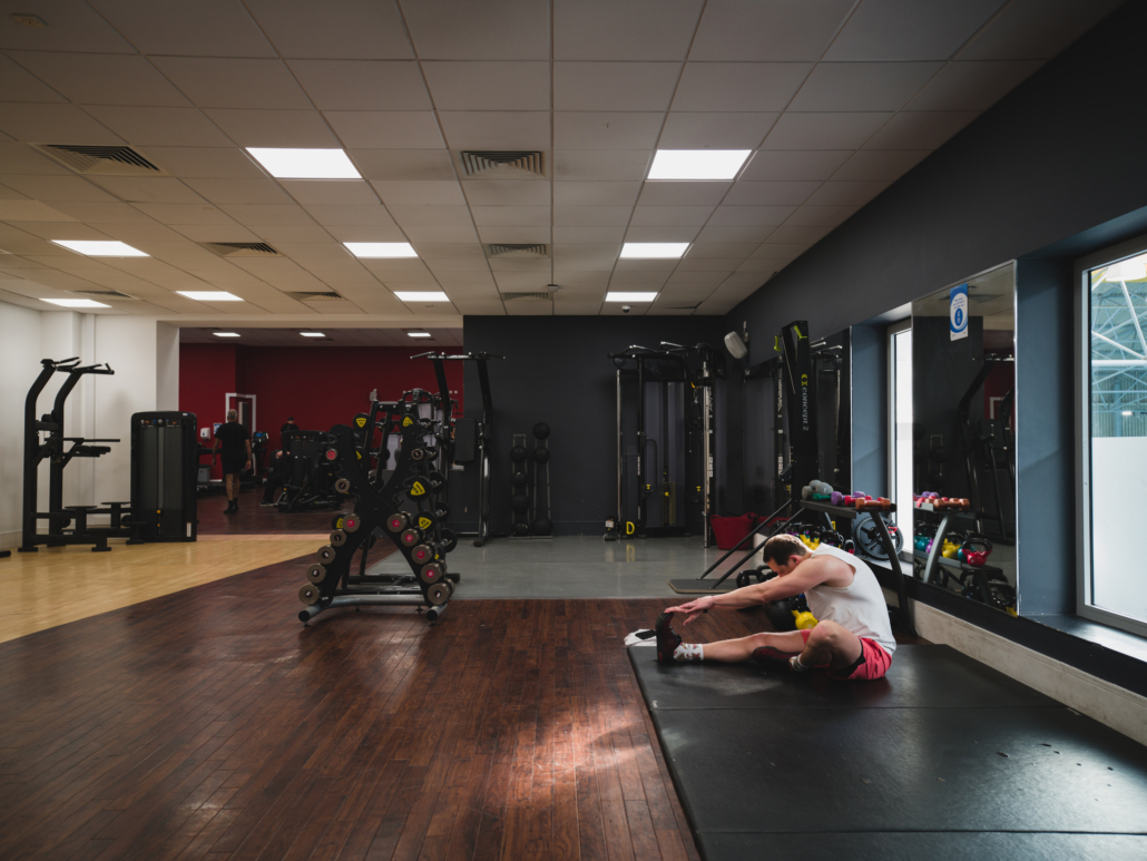 A person in pink shorts is stretching on a black mat in a gym, surrounded by various exercise equipment and weights.