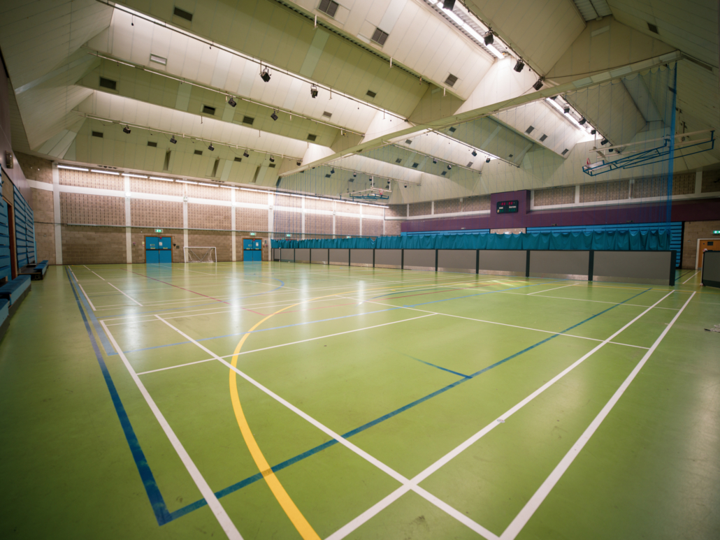 Spacious indoor sports hall with high ceiling, green flooring, blue markings, basketball hoops, and a scoreboard. No people present.