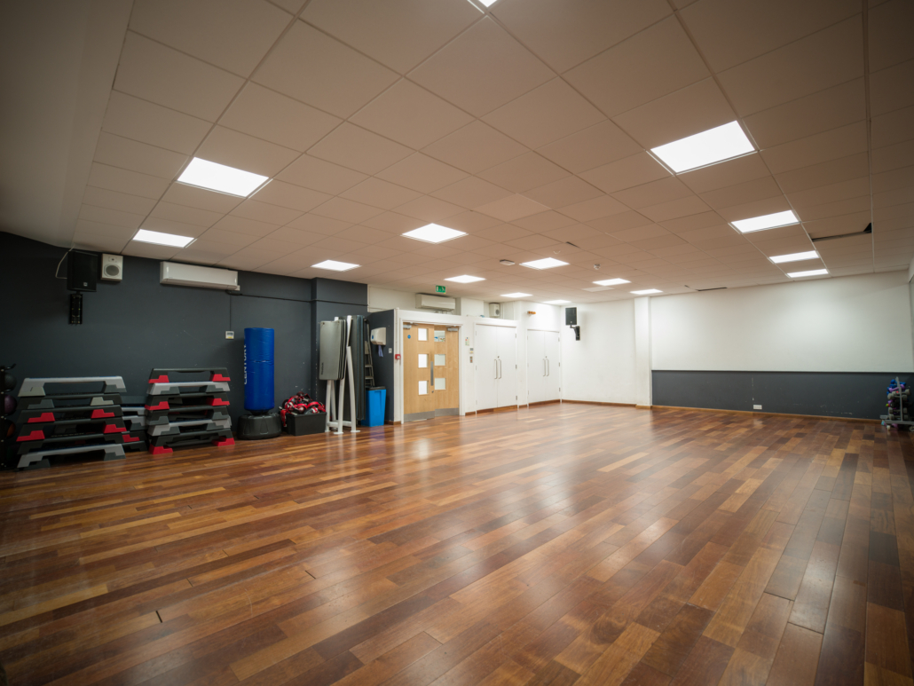 Spacious room with wooden flooring, stacked exercise equipment, a punching bag, and bright ceiling lights, ideal for fitness classes or workouts.